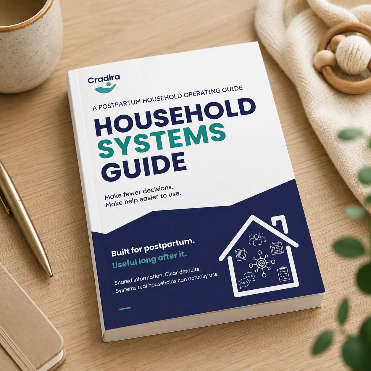 Paperback copy of the Cradira Household Systems Guide resting on a wooden table beside a mug, pen and soft baby items.